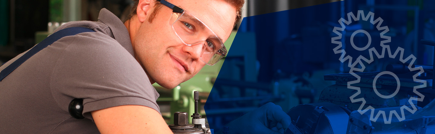 Header: Ein Maschinenbauer ist mit einer Schutzbrille in die Kamera lächelnd über eine Werkbank gebeugt. 